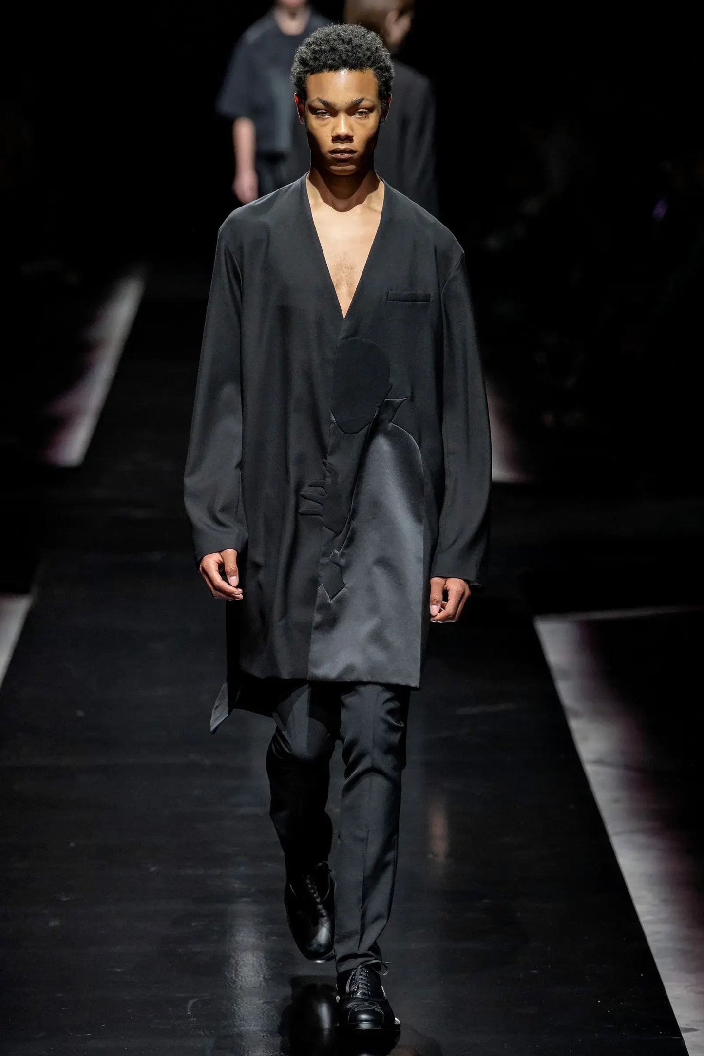 Tasoni black wool coat with asymmetric collarless design and Dracula motive print, worn by a male model on a runway.