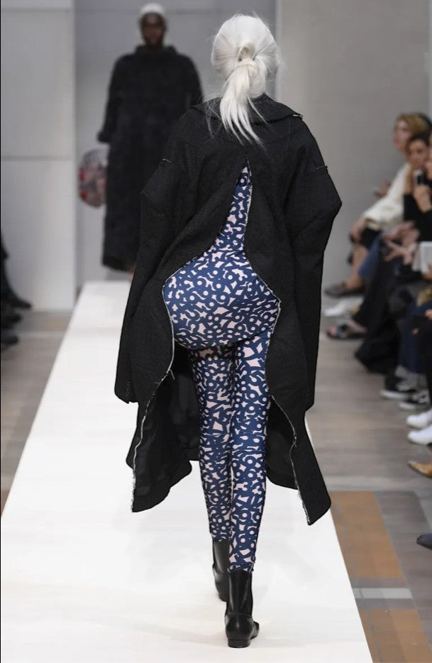 Tasoni Comme des Garcons D-Pattern leggings in navy and pink, worn under a black coat on a runway.