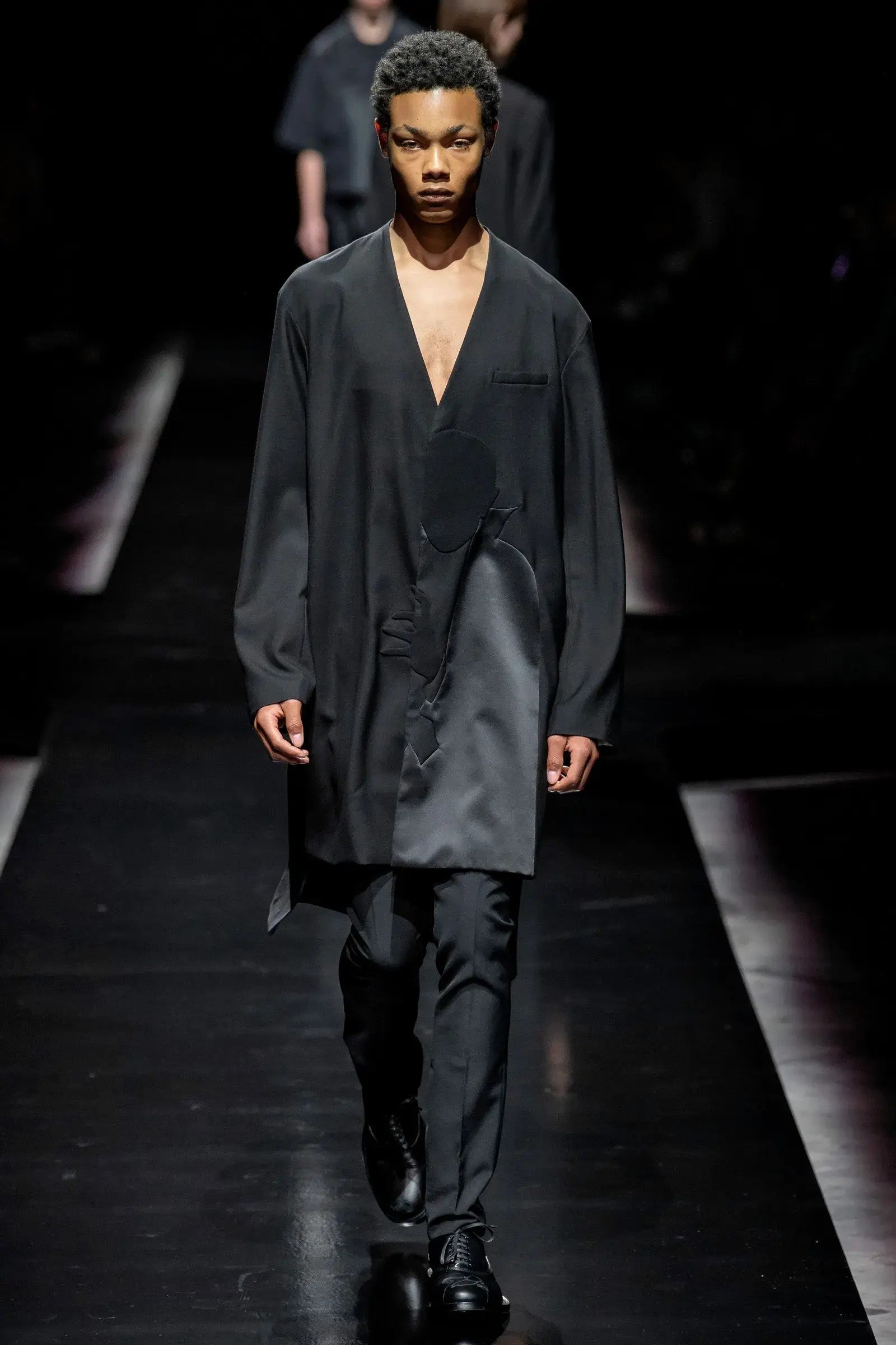 Tasoni black wool coat with asymmetric collarless design and Dracula motive print, worn by a male model on a runway.