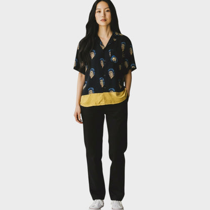 Tasoni black silk blend short sleeve shirt with a repeating David Bowie print and a mustard yellow hem. Model wears black trousers and white sneakers.