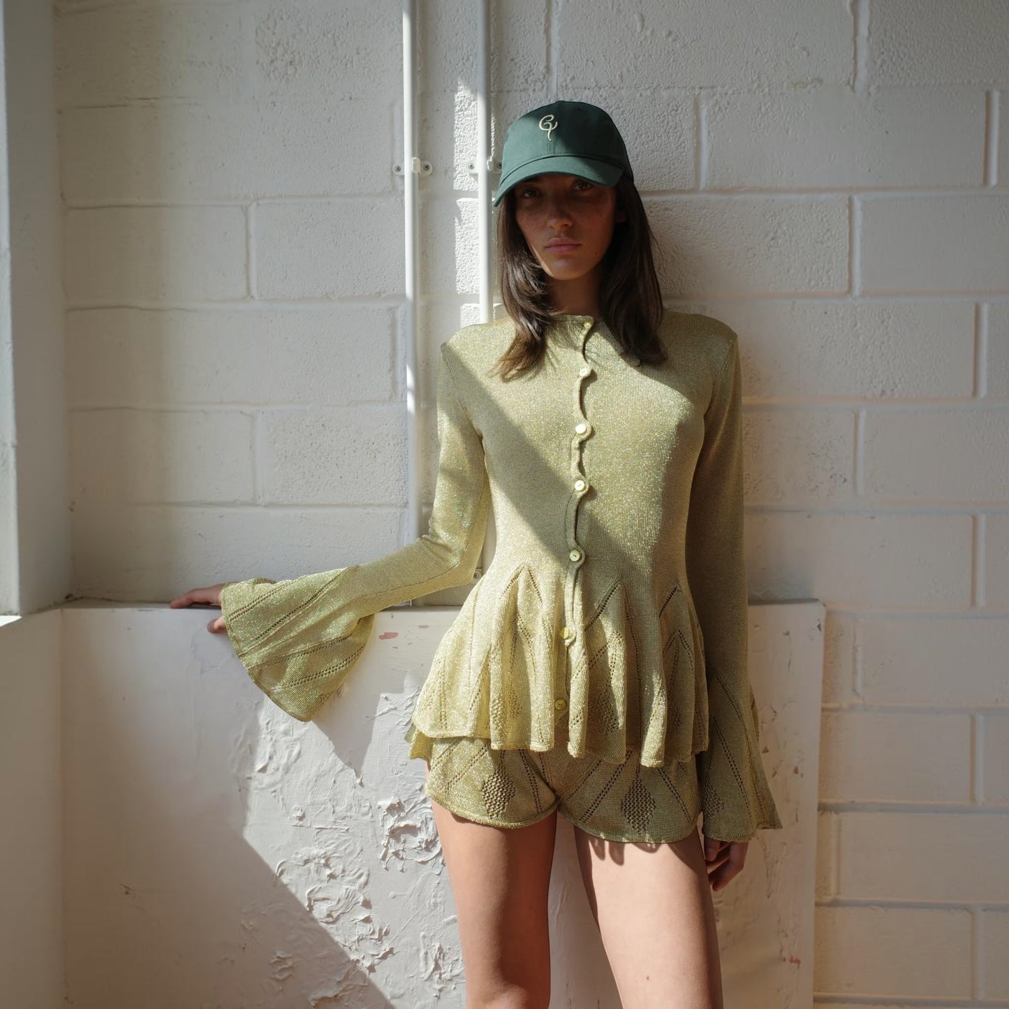 Model wearing Tasoni gold knitted pointelle godet shorts and matching flared-sleeve cardigan against a white wall.