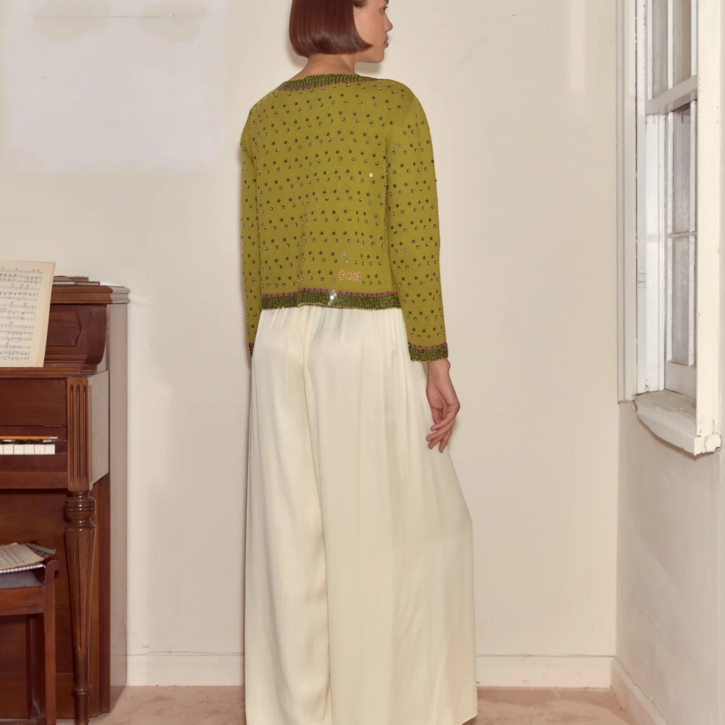 Tasoni green knit cardigan with fairy floss sequins, crew neck, and beaded trim. Model wears cream wide-leg pants.