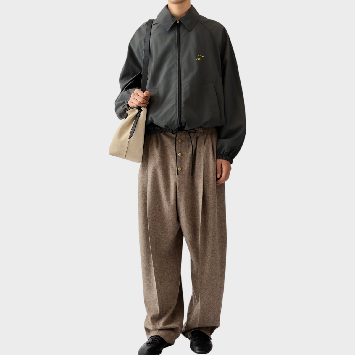 Tasoni Jacquemus Le Coupe-Vent charcoal grey windbreaker jacket with a small yellow logo, worn with brown trousers.