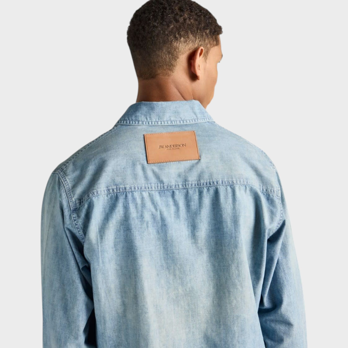 Back view of a man wearing a Tasoni J.W.Anderson pale blue cotton denim shirt with a tan leather logo patch.