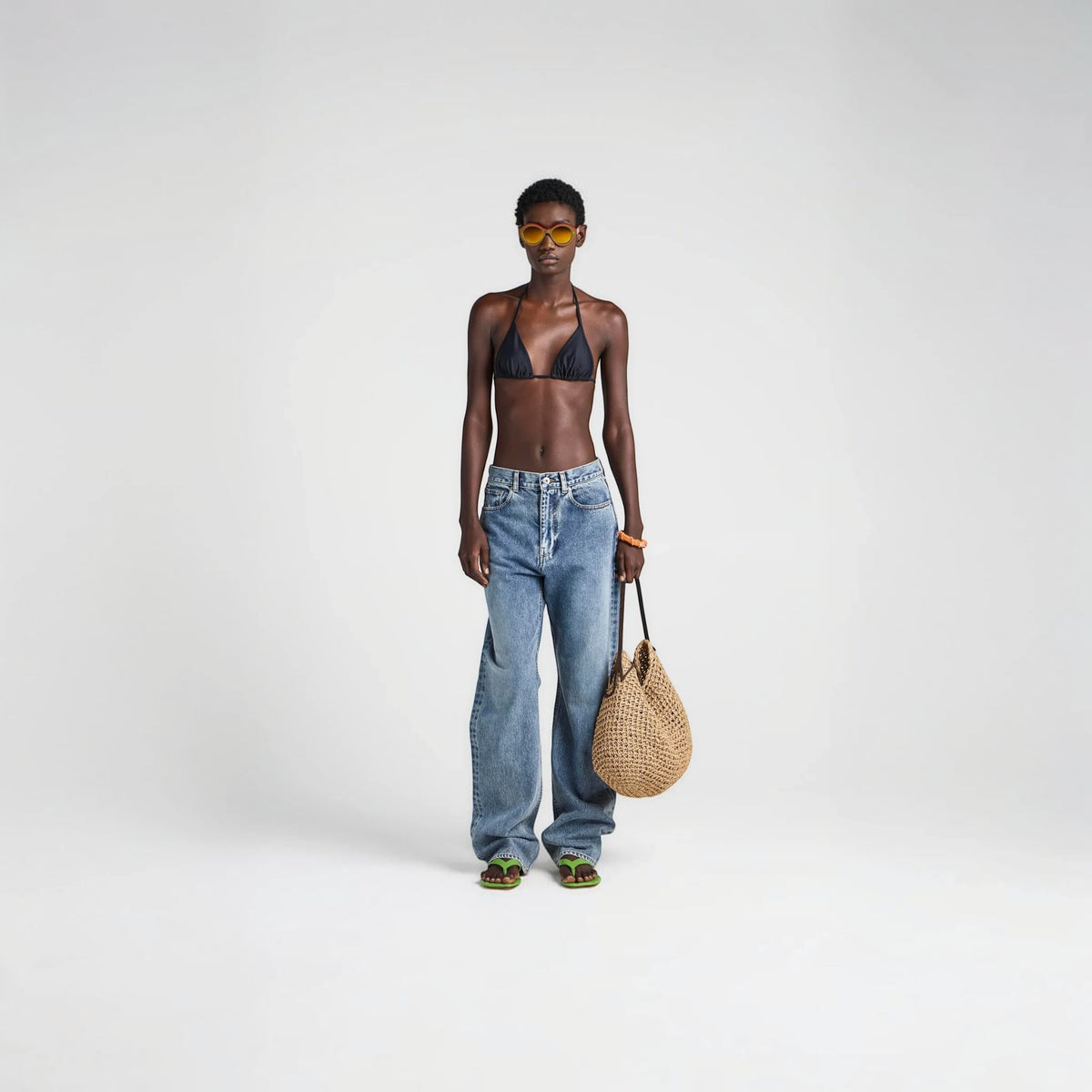Model wearing a black JW Anderson triangle bikini top from Tasoni, styled with blue jeans and a woven straw bag.