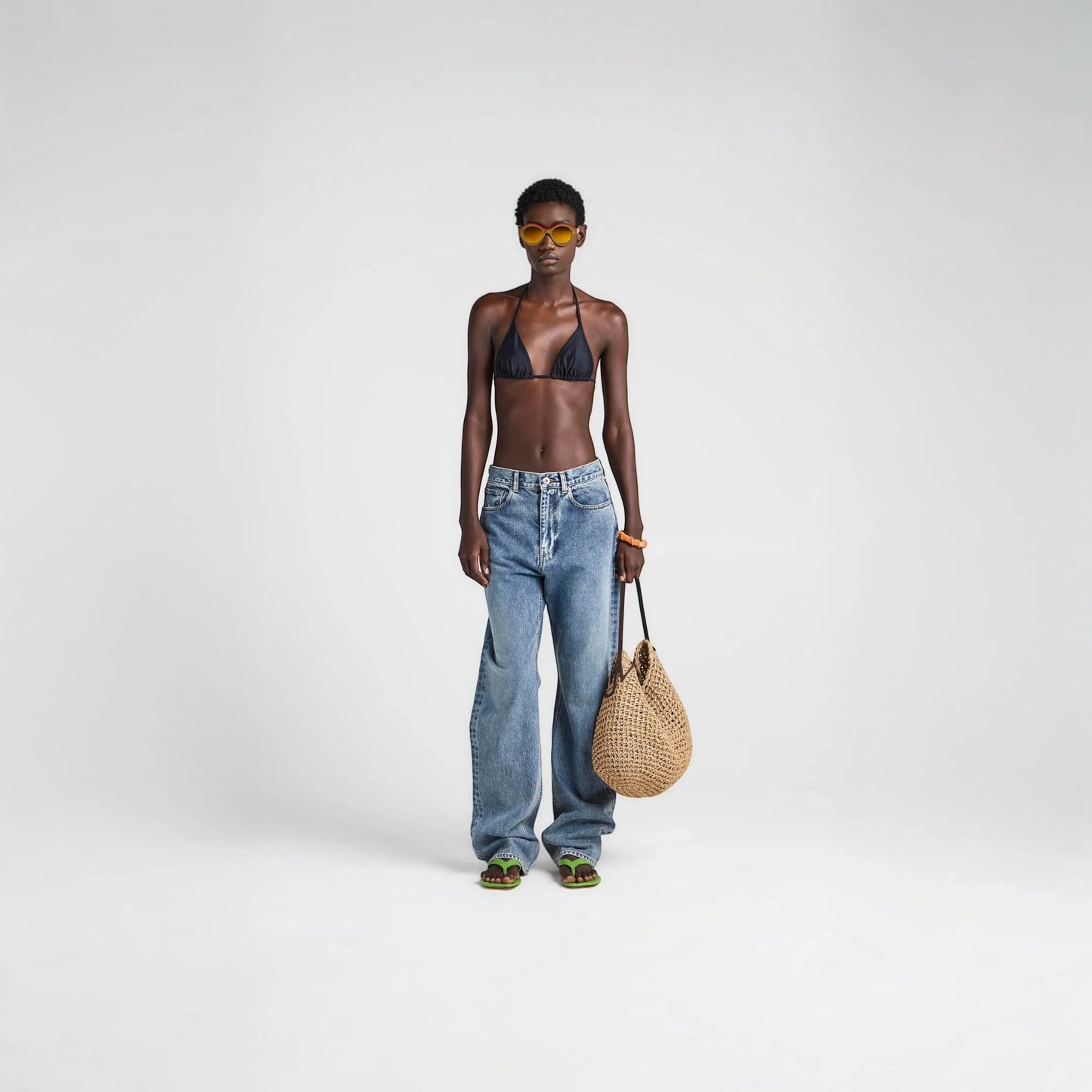 Model wearing a black JW Anderson triangle bikini top from Tasoni, styled with blue jeans and a woven straw bag.