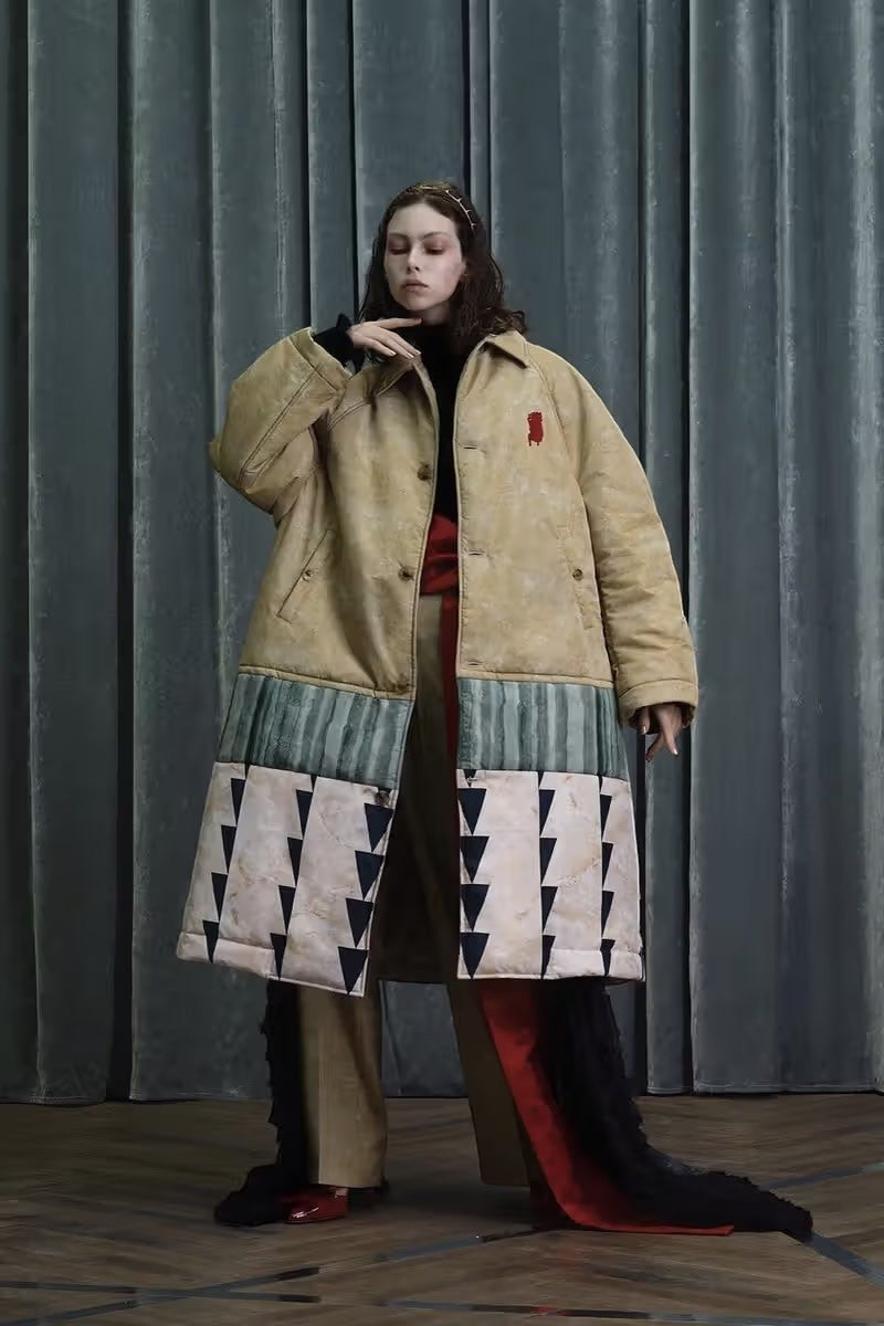 Tasoni oversized beige padded coat with a suspirium print, featuring a red embroidered logo and a multi-panelled hem with abstract patterns.