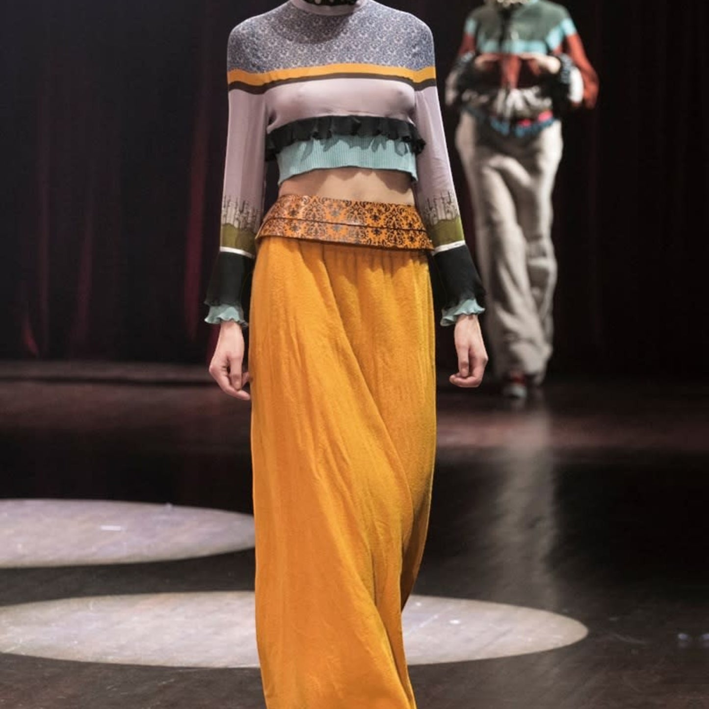 Tasoni model wears a long-sleeved, multi-colored patterned silk top with ruffled trim and a high neck, paired with an orange maxi skirt.