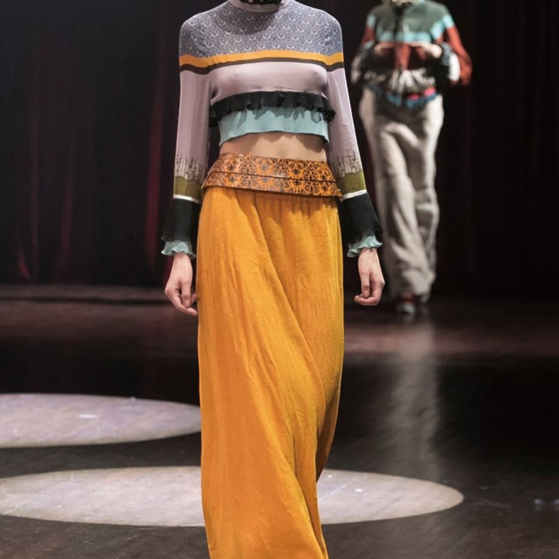 Tasoni model wears a long-sleeved, multi-colored patterned silk top with ruffled trim and a high neck, paired with an orange maxi skirt.